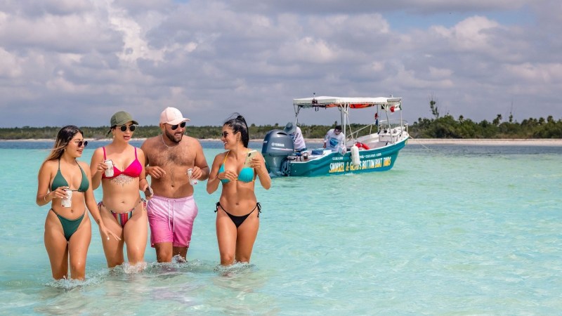 Four people in swimwear walking in shallow turquoise water, with a boat and beach in the background.