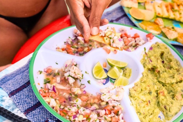 Person holding chip with ceviche near platter of guacamole and lime wedges.