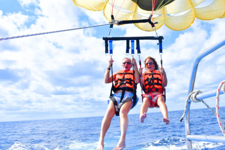 Two people parasailing with a yellow smiley face parachute over the ocean.