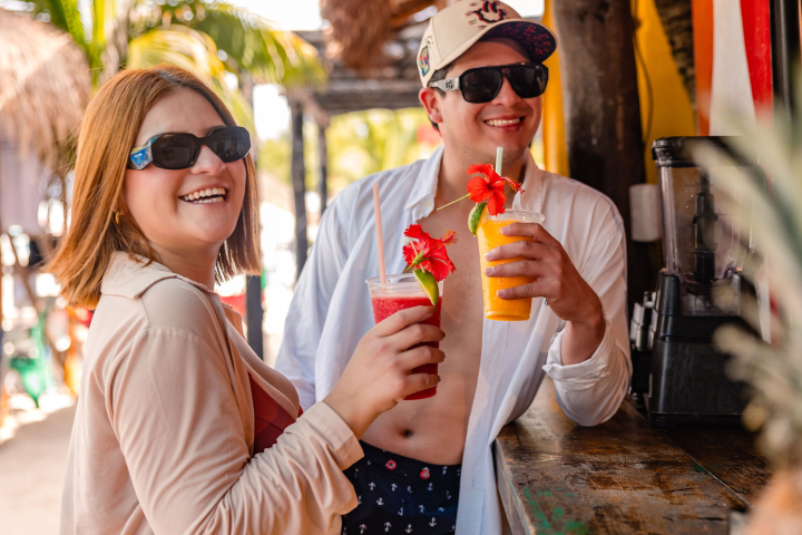 Two people in sunglasses enjoy tropical drinks at a beach bar.