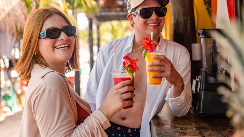 Two people in sunglasses enjoy tropical drinks at a beach bar.
