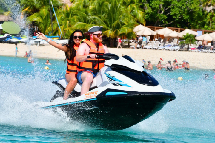Two people on a jet ski in tropical waters wearing life vests, with palm trees in the background.