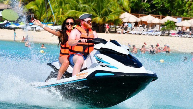 Two people on a jet ski in tropical waters wearing life vests, with palm trees in the background.