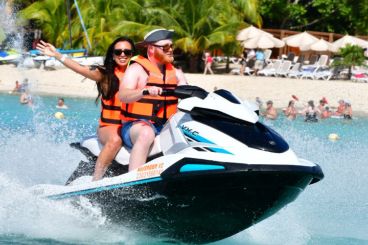 Two people on a jet ski in the ocean, wearing life vests, with a beach and palm trees in the background.