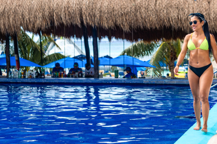Woman in bikini standing by a pool under a thatched roof with palm trees and blue umbrellas in background.