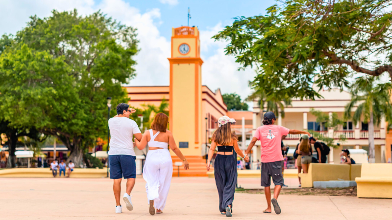 Four people walk toward a clock tower in a sunny plaza with trees and benches.