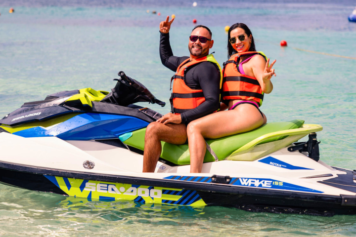 Two people in life jackets on a Sea-Doo jet ski, smiling and giving peace signs.
