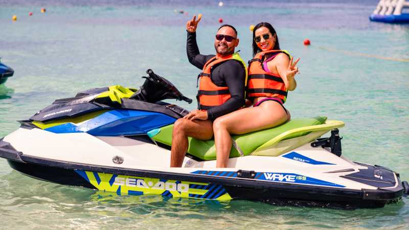 Two people in life jackets on a Sea-Doo jet ski, smiling and giving peace signs.