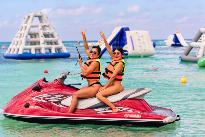 Two people on a red jet ski in clear blue water with inflatables in the background.