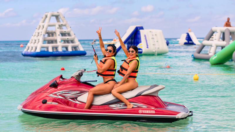 Two people on a red jet ski in clear blue water with inflatables in the background.