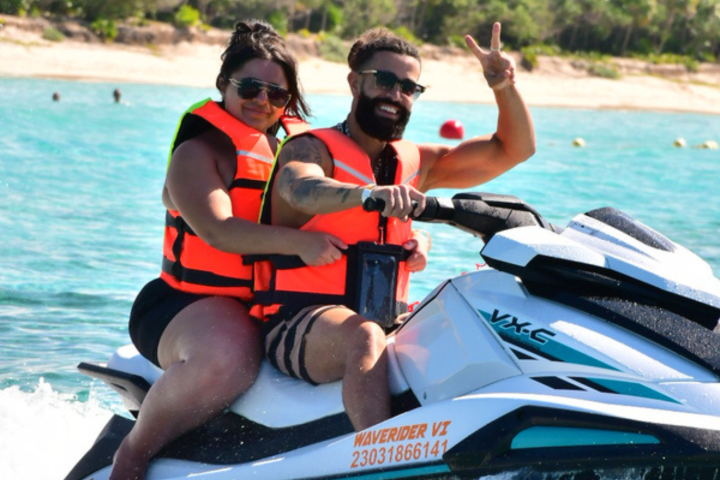 Two people on a jet ski in life vests, showing a peace sign, with beach in background.