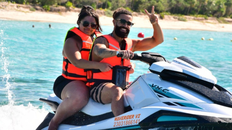 Two people on a jet ski in life vests, showing a peace sign, with beach in background.