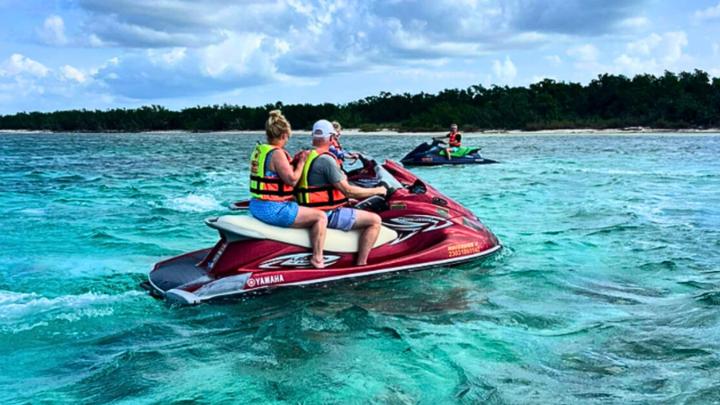 People riding jet skis in clear turquoise water near a forested shoreline.