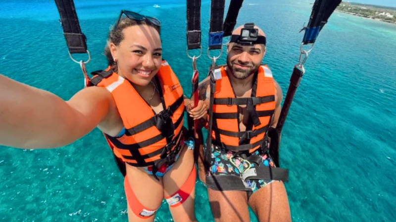 Two people parasailing over clear blue water, wearing orange life vests.