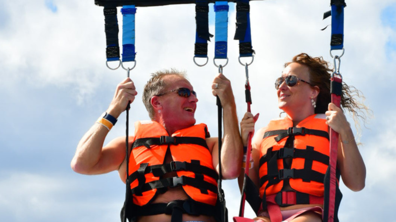 Two people parasailing with orange life vests, smiling at each other.