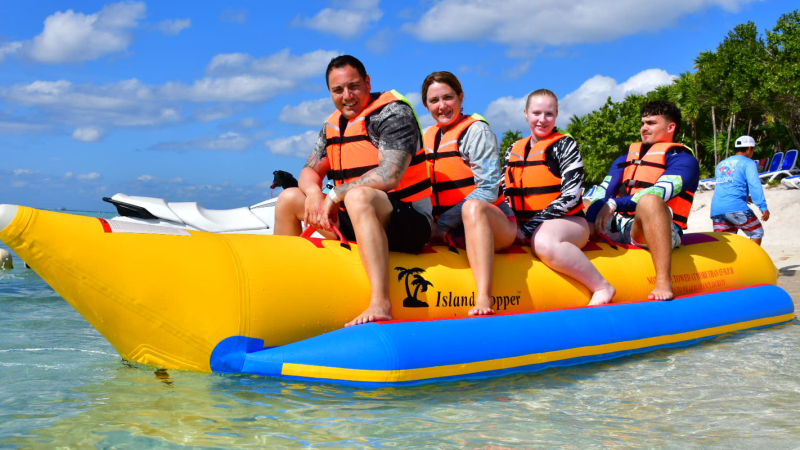 Four people on a yellow inflatable banana boat on the water, wearing life jackets.